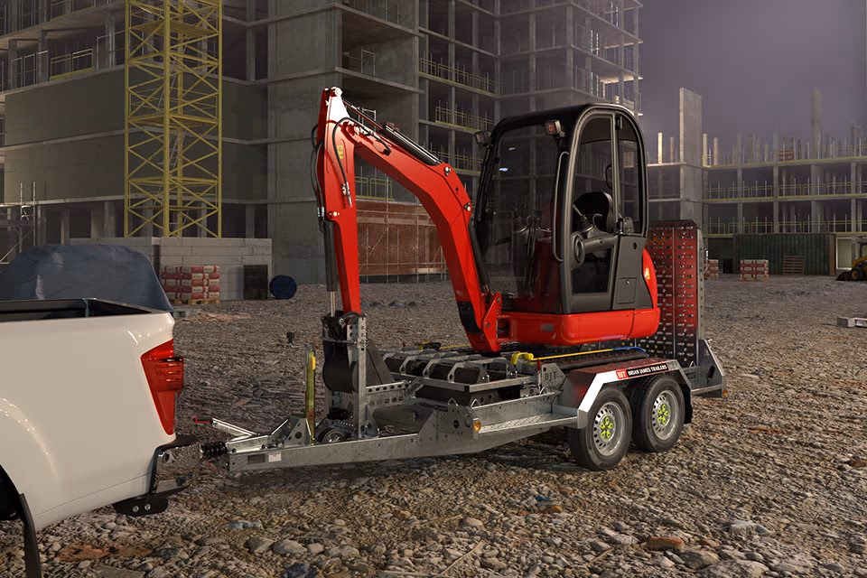 Miniescavatore caricato su rimorchio Brian James Digger Plant agganciato a pickup, in un cantiere edile.