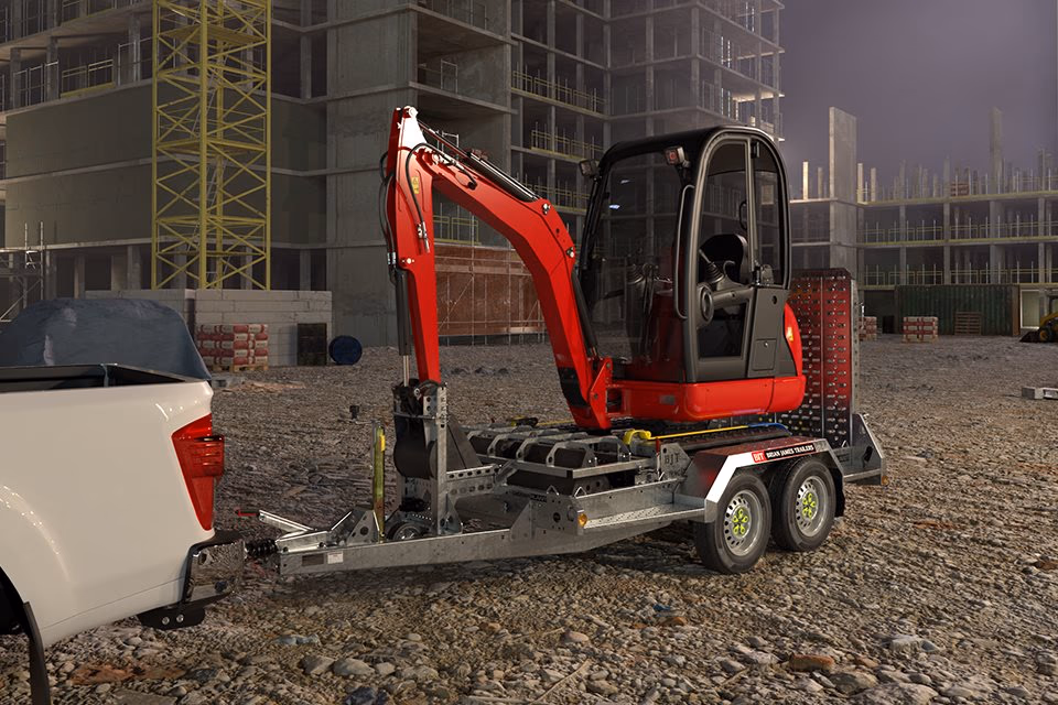 Miniescavatore caricato su rimorchio Brian James Digger Plant agganciato a pickup, in un cantiere edile.